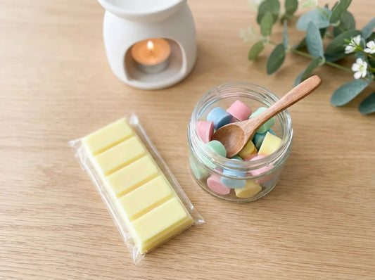 Wax melt snap bar and an open tub of wax melt scoopies beside a white tealight burner on a light wooden table in soft daylight.